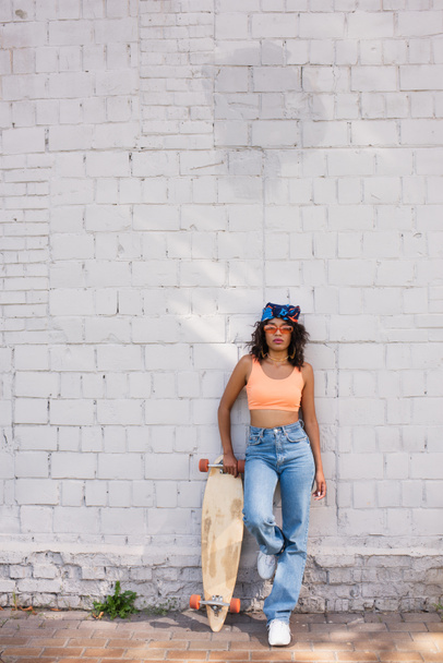 κομψή αφρικάνικη αμερικανίδα γυναίκα με γυαλιά ηλίου και μαντίλα κρατώντας longboard κοντά σε τοίχο από τούβλα - Φωτογραφία, εικόνα