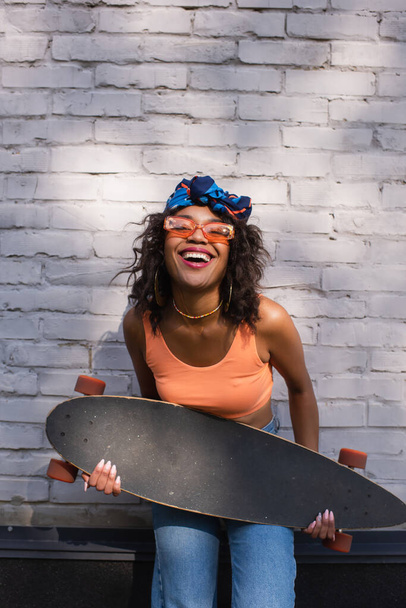 šťastný mladý africký americký žena v sluneční brýle a šátek držení longboard poblíž cihlové zdi - Fotografie, Obrázek