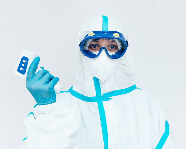 Un médico con traje médico protector y máscara sostiene un termómetro infrarrojo moderno en su mano. - Foto, Imagen