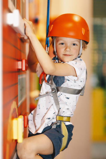 Indoor-Kletterkurs für Kinder. Kleiner Junge mit rotem Helm klettert die Wand im Boulderzentrum hinauf. Schulkind lächelt in die Kamera und hat Spaß auf Indoor-Spielplatz für Kinder - Foto, Bild