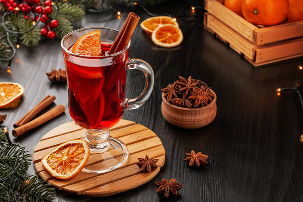 A glass of mulled wine with cinnamon sticks, orange slices and anise stars on a wooden stand on a dark background with fir branches and a garland. The atmosphere of comfort, Christmas, holiday. Advertising, banner. Horizontal photo. - Foto, Imagem