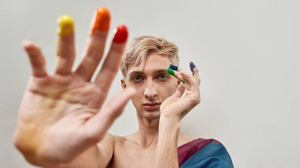 Young transgender with fingers in colorful rainbow lgbtqi paint looking at camera while posing isolated over light background - Foto, afbeelding