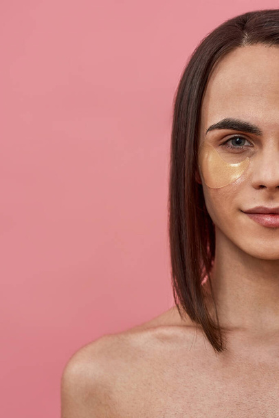 Half face closeup of shirtless young transgender with dark brown and applied under eye gel patches looking at camera, taking care of skin, posing isolated over pink background - Foto, afbeelding