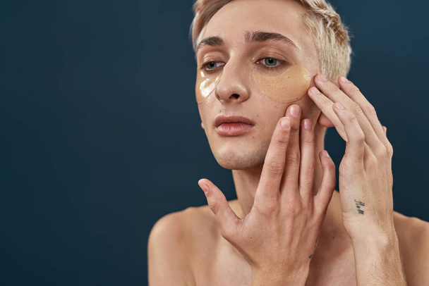 Primer plano retrato de jóvenes transgénero aplicando bajo parches de gel ocular, cuidando la piel mientras posan aislados sobre un fondo oscuro - Foto, Imagen