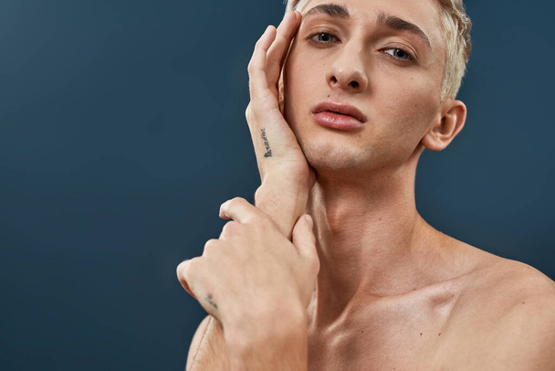 Portrait of androgynous transgender young man with makeup and light hair touching his face and looking at camera while posing isolated over dark blue background - Photo, image