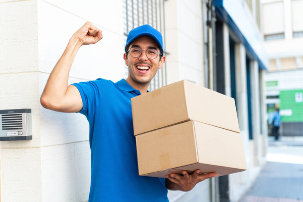 Young delivery man at outdoors holding boxes and celebrating a victory - Фото, изображение