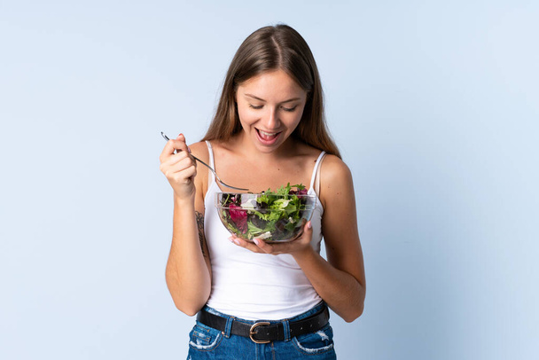 Genç Litvanyalı bir kadın mavi arka planda izole edilmiş ve elinde mutlu bir ifadeyle bir kase salata tutuyor. - Fotoğraf, Görsel