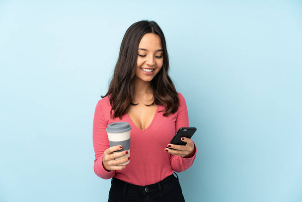 Jeune femme métisse isolée sur fond bleu tenant du café à emporter et un mobile - Photo, image