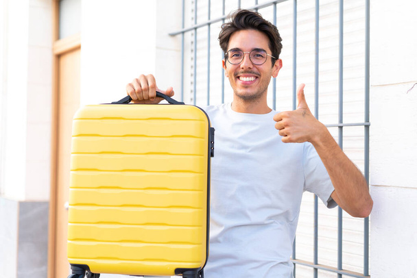 Nuori valkoihoinen mies ulkona lomalla matkalaukku ja peukalo pystyssä - Valokuva, kuva