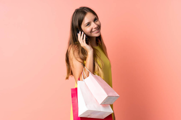 Молодая литовская женщина изолирована на розовом фоне, держит сумки с покупками и звонит другу на мобильный телефон - Фото, изображение