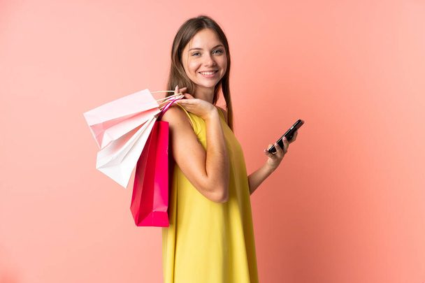 Young Lithuanian woman isolated on pink background holding shopping bags and writing a message with her cell phone to a friend - Foto, immagini