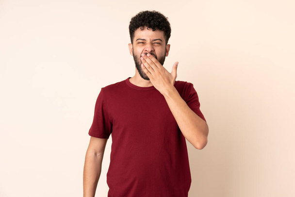 Jovem marroquino isolado no fundo bege bocejando e cobrindo ampla boca aberta com a mão - Foto, Imagem