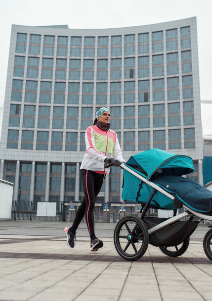 Active young mom in sportswear training and pushing stroller with a child in the city on a cold autumn day - Foto, Imagem