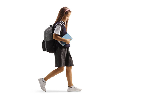 Full length profile shot of a sad female pupil in a school uniform walking and holding a book isolated on white background - Foto, afbeelding
