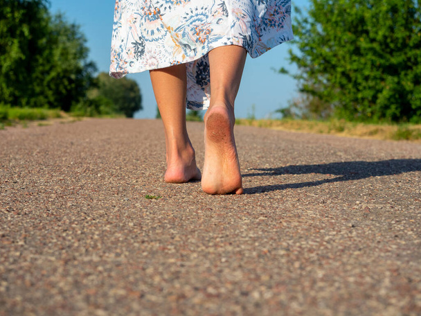 Una donna vestita con un vestito cammina scalza sull'asfalto. Vista posteriore. Il concetto di viaggio, ricreazione, vacanza, libertà - Foto, immagini