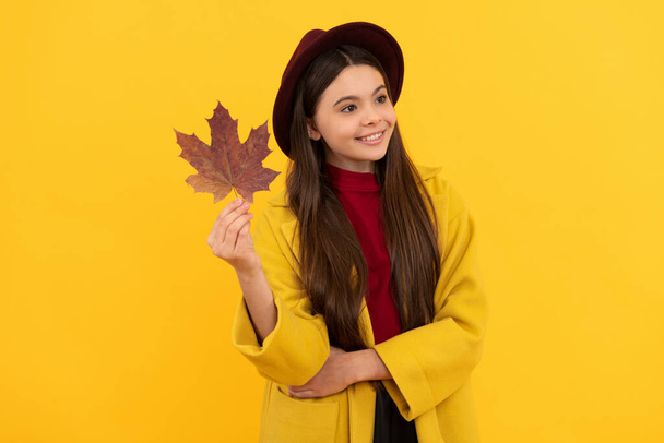 smiling kid in hat and coat with autumn maple leaf on yellow background, season - Photo, Image