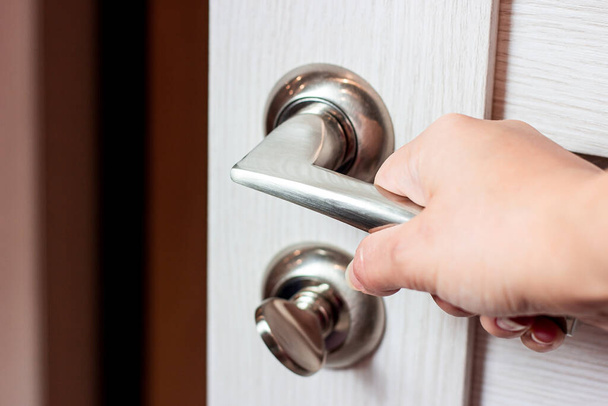 Caucasian woman hand pushes the modern metal doorknob on the white wooden door in the house to open it. - Photo, Image