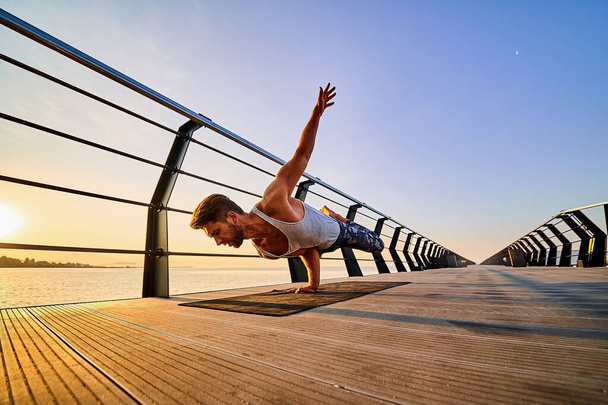 Fit man doing a one hand stand while practicing yoga alone near the ocean against sky at dusk or dawn - Foto, afbeelding