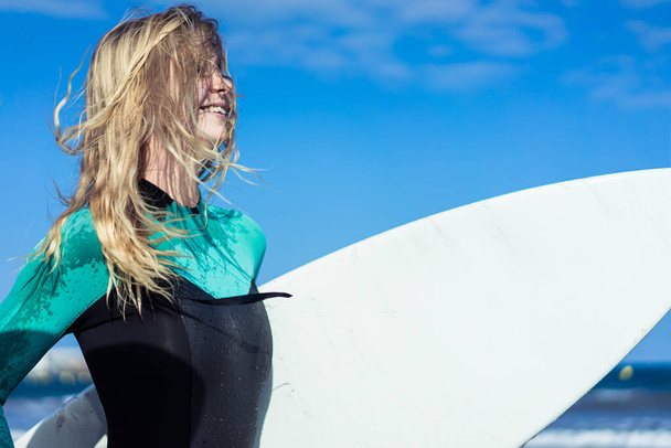 Young sexy girl with long blonde hairs, smiling and jogging with surf board on the beach in a summer day. Background blue sky. Copy space. - Photo, image