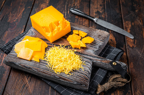 Geraspte en in blokjes gesneden Cheddar Cheese op een houten snijplank. Donker houten achtergrond. Bovenaanzicht - Foto, afbeelding
