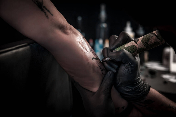 Una toma horizontal de un artista del tatuaje haciendo un tatuaje a una mujer en su brazo, el artista del tatuaje está muy concentrado operando la máquina de tatuaje - Foto, Imagen