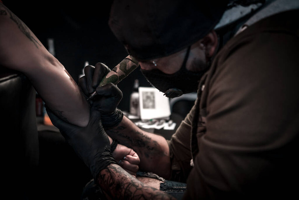 Una toma horizontal de un artista del tatuaje haciendo un tatuaje a una mujer en su brazo, el artista del tatuaje está muy concentrado operando la máquina de tatuaje - Foto, Imagen