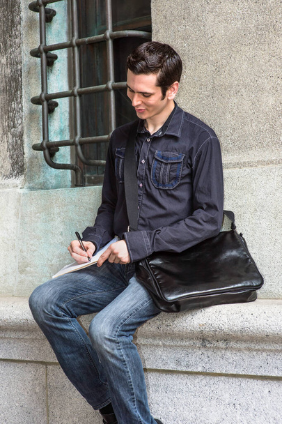Llevando una bolsa de cuero y sentado junto a una ventana de estilo antiguo, un joven estudiante guapo está tomando una nota afuera. - Foto, imagen