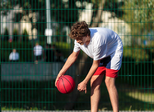 Adolescent mignon joue au basket sur une aire de jeux de la ville. Un garçon tient un ballon de basket dans ses mains dehors. Vie active, passe temps, sport pour enfants - Photo, image