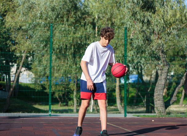 かわいいティーンエイジャーは、都市の遊び場でバスケットボールをします。少年は外でバスケットボールボールを手に持っている。子供のためのアクティブな生活、趣味、スポーツ - 写真・画像