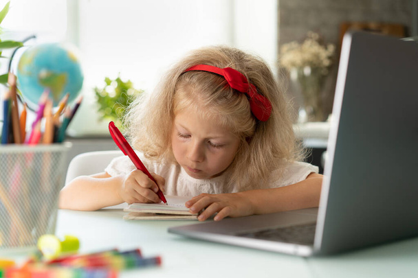 μικρό κορίτσι κάνει μαθήματα στο σπίτι σε ένα φορητό υπολογιστή, γράφει με ένα στυλό σε ένα σημειωματάριο. - Φωτογραφία, εικόνα