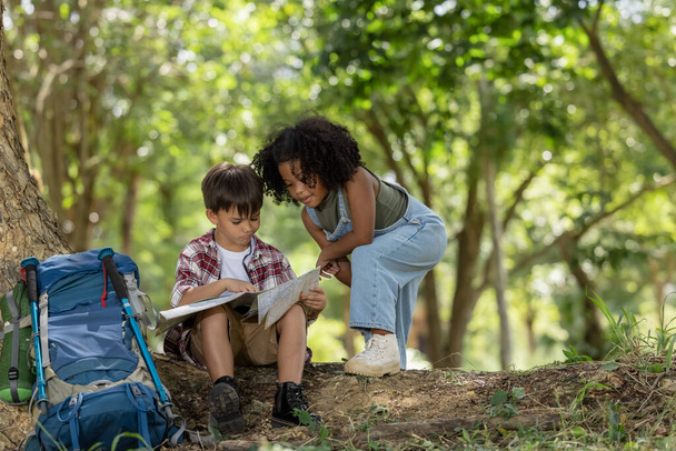 African American ethnicity boy and girl children wearing plaid shirt and backpack sitting at tree base talking and looking down on the map travel on hand in the park background hiking adventure summer holiday concept - Fotoğraf, Görsel