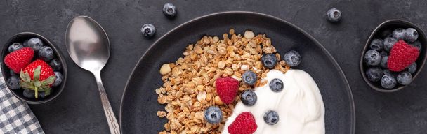Horizontal banner with homemade granola, yogurt and fresh berries in black ceramic plate on dark stone background. Healthy vegan breakfast - Фото, изображение
