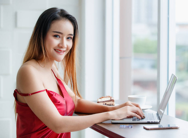 Mooi jong gesneden Aziatische sexy meisje op rood verleidelijke jurk mooie glimlach naar andere als ze geniet van het werk op laptop in heldere ochtend met ontbijt van een stuk taart en een kopje koffie. - Foto, afbeelding