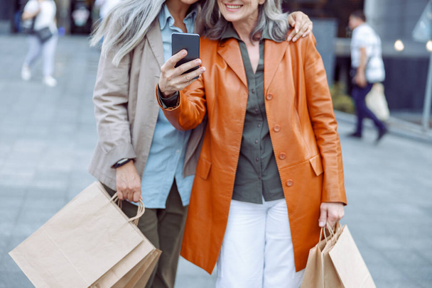 Senior κυρία με δερμάτινο μπουφάν και σύντροφος με ψώνια λάβει selfie στο σύγχρονο δρόμο της πόλης - Φωτογραφία, εικόνα