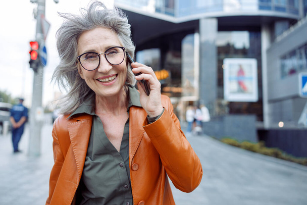 Happy grey haired senior woman with glasses talks on mobile phone on city street - Photo, Image