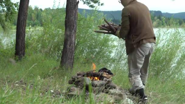 Muž v lese na břehu řeky nese náruč plnou suchého dřeva a dává je do ohně. - Záběry, video