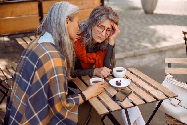 Depressziós érett hölgy szemüveggel és gondos barát si utcai kávézóban ősszel nap - Fotó, kép