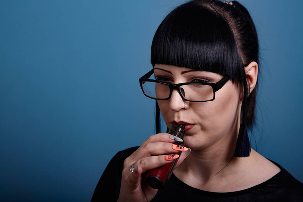 A beautiful girl with dark hair and glasses exquisitely smokes an electronic cigarette on a dark background - Photo, Image