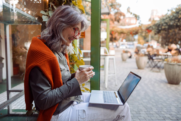 Gyönyörű idős nő használ videochat laptopon ül padon közelében kávézó - Fotó, kép
