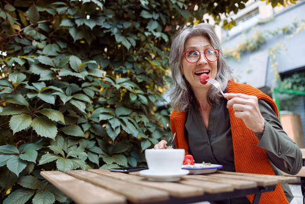 Весела старша жінка з окулярами їсть полуничний десерт, сидячи за столом на відкритій терасі кафе
 - Фото, зображення