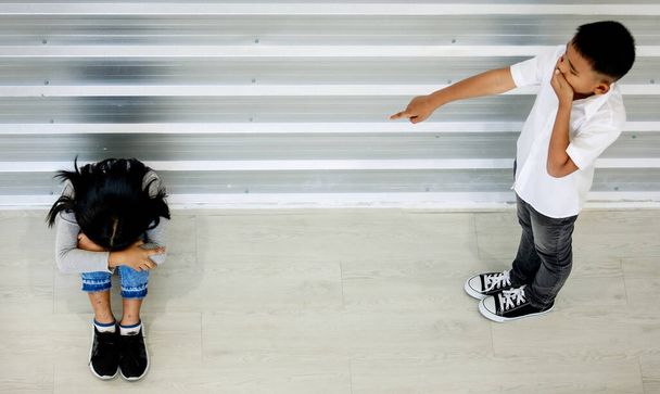 Top view shot z Azji młody niesforny hałaśliwy szalony chłopiec stojący śmiech obwinia wskazując nękanie na małe zdenerwowany smutny dziewczyna siedzi na podłodze pokrywa twarz między kolanami płacze samotnie w szkole podstawowej. - Zdjęcie, obraz