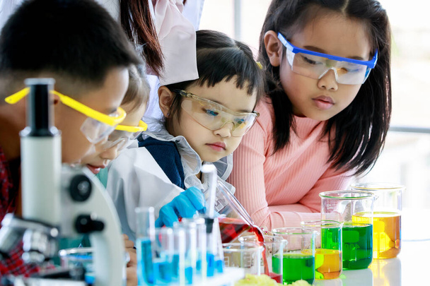 アジア系の好奇心旺盛な就学前の女の子が先生を観察し、ガラス製のビーカーから赤い試薬を注ぐことは、顕微鏡や若い友人と機器を用いた実験室のテーブルで化学実験を行います. - 写真・画像