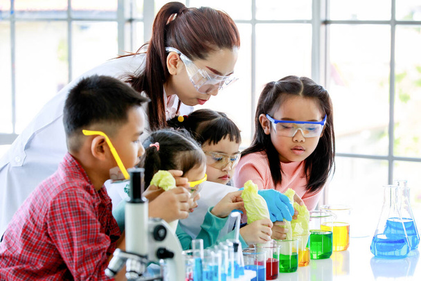 Cam deney şişesinden kırmızı reçel döken öğretmeni gözlemleyen Asyalı küçük meraklı anaokulu kızı genç arkadaşlarıyla birlikte mikroskop ve ekipmanla laboratuvar masasında kimyasal deney yapıyor.. - Fotoğraf, Görsel