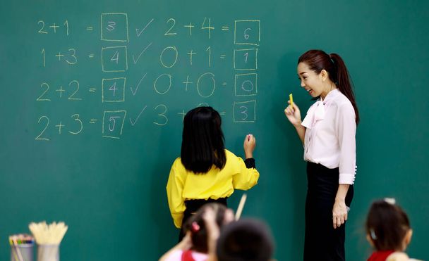 Πορτρέτο πλάνο της ασιατικής όμορφη γυναίκα μαθηματικός στέκεται χαμογελώντας ευθυμία, ενώ λίγο έξυπνη μαθήτρια επίλυση εξισώσεις μαθηματικά μαθηματικό σε πίνακα μπροστά από συμμαθητές στην τάξη. - Φωτογραφία, εικόνα