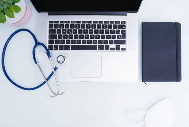 Stethoscope over laptop and notebook, plant and mask on white table with copy space. - Foto, imagen