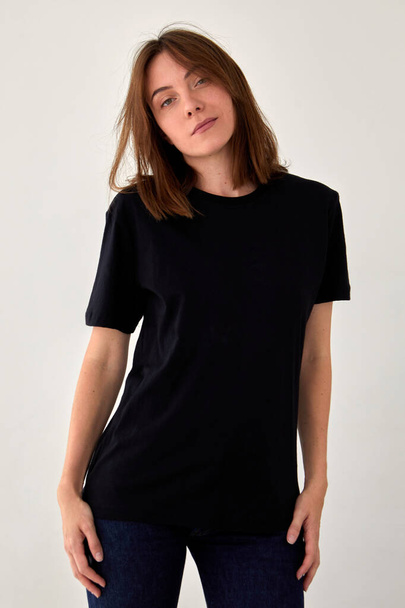 Gelassene junge Frau trägt lässiges schwarzes T-Shirt und schaut in die Kamera auf weißem Hintergrund im Studio - Foto, Bild
