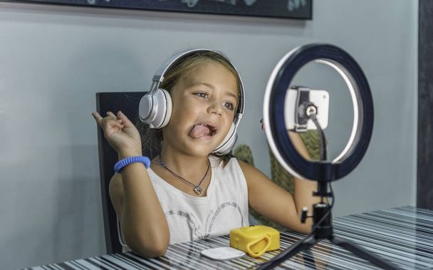 Caucasian little girl making faces while making a video with cell phone and headphones at home. Concept of new technologies - Fotografie, Obrázek