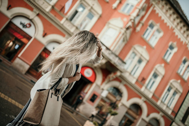 Μια ξανθιά γυναίκα με μια τσάντα περπατώντας στο δρόμο με ένα κόκκινο κτίριο στο παρασκήνιο. - Φωτογραφία, εικόνα