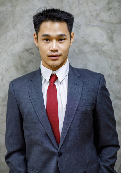 Joven hombre asiático de pelo corto con camisa blanca, corbata roja, traje gris oscuro, de pie ligeramente sonriente, mirando la cámara, foto de fotos en el estudio con un fondo loft - Foto, Imagen