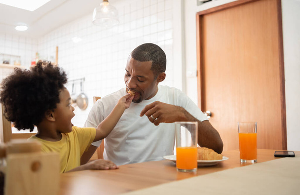 Happy African American Father and little son eating Croissant and drinking juice for breakfast in dining room together. - 写真・画像